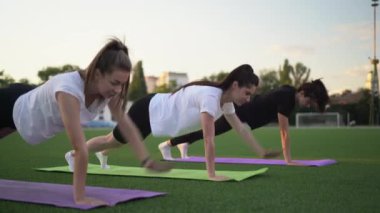 Bir grup kadın stadyumda antrenman yapıyor, tahta bir pozisyonda duruyorlar, bacaklarını kaldırıyorlar. Açık hava fitness konsepti