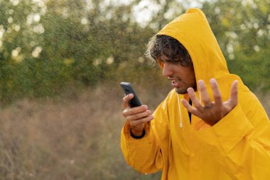 Yağmurluk giymiş kızgın bir adam soğuk yağmurda ayakta dikilirken telefona bağırıyor. Kötü hava