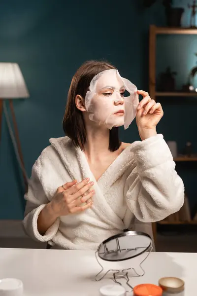 Self-care concept. Skin care beauty routine. Portrait of young beautiful woman wearing white bathrobe chilling in bedroom and removing facial sheet mask, doing beauty treatment. Copy space