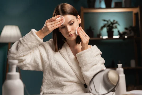 Self-care concept. Skin care beauty routine. Portrait of young beautiful woman wearing white bathrobe chilling in bedroom and cleansing her face with pads, doing beauty treatment and relaxing at home.