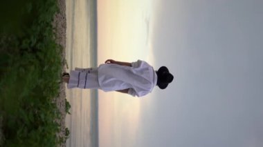 Deniz kıyısında duran şapkalı genç bir kadın ufkun ötesindeki mesafeye bulutlu bir gökyüzünün arka planına bakıyor. Yaz tatili kavramı. Dikey video