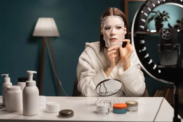 Woman in white bathrobe records video tutorial at home with sheet mask on her face shoots broadcast live video to social network. Blogging, skin care and beauty concept. Copy space