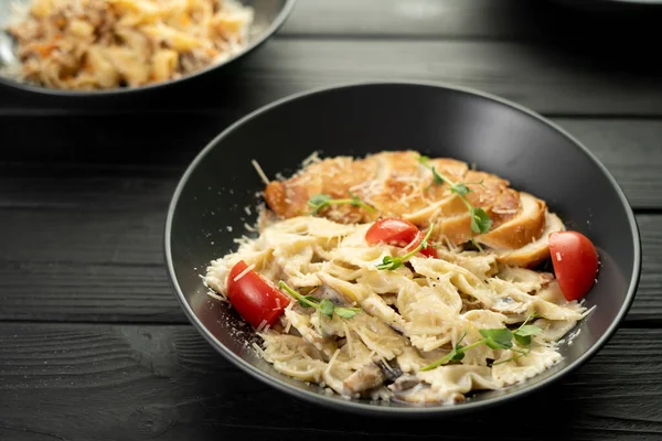 Farfalle with mushrooms, chicken fillet and parmesan in cream sauce in a black plate on the wooden table. Traditional Italian pasta on a dark culinary background. Copy space
