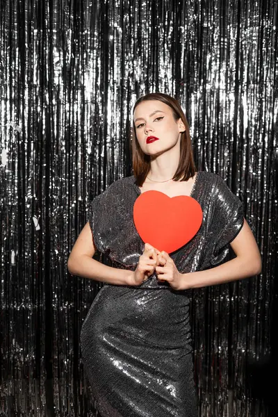 Young woman in shining dress hold in hands red heart paper shape on gray sparkling tinsel background. Valentines Day concept