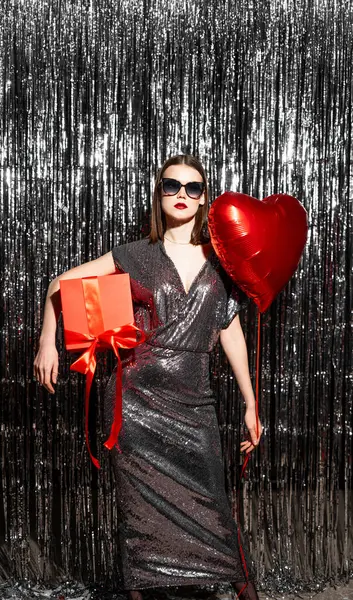 Beautiful young woman in shining glamour dress with heart shape air balloon and gift box on tinsel sparkling background. Woman on Valentine's Day. Symbol of love. Copy space