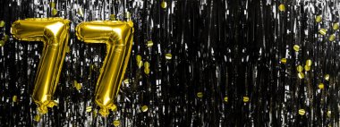 Siyah süslemenin arka planında 77 numaralı altın folyo balonu. Doğum günü tebrik kartı, 77 nolu yazı. Yıldönümü etkinliği. Pankart. Boşluğu kopyala.