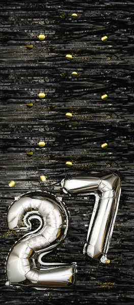Silver foil balloon number 27 on a background of black tinsel decoration with golden confetti. Birthday greeting card, inscription twenty-seven. Anniversary event. Vertical Banner. copy space.