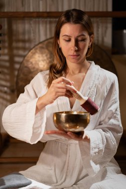 Kadın yoga minderinin yanında otururken Tibet şarkı kasesi kullanıyor. Boşluğu kopyala