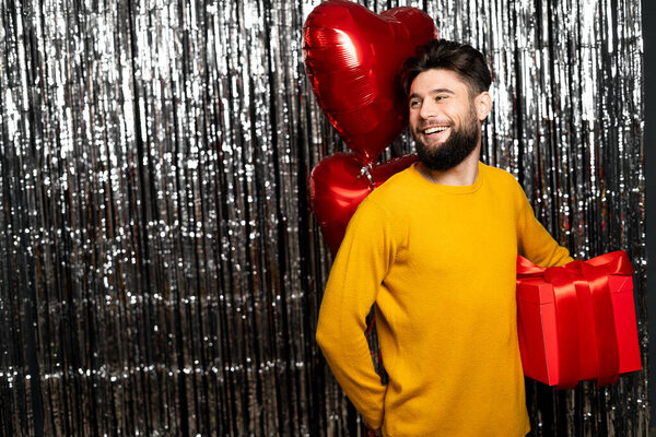 Handsome young man holding red gift and heart shaped balloons, smiling while standing on tinsel background. St Valentine's Day concept. Copy space