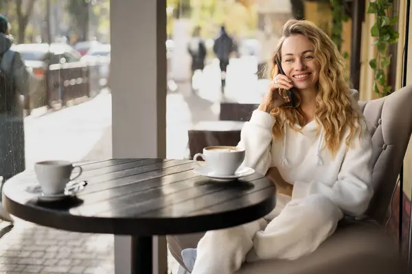 Happy blonde woman speaking with her friend by phone while relaxing in cozy coffee shop, talking on smartphone while sitting in cafe during break. Copy space