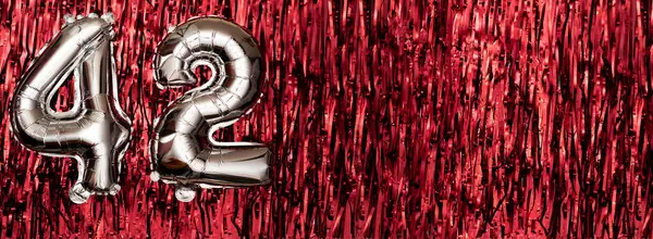 Silver foil balloon number 42 on a background of red tinsel decoration. Birthday greeting card, inscription forty-two. Anniversary event. Banner. copy space.