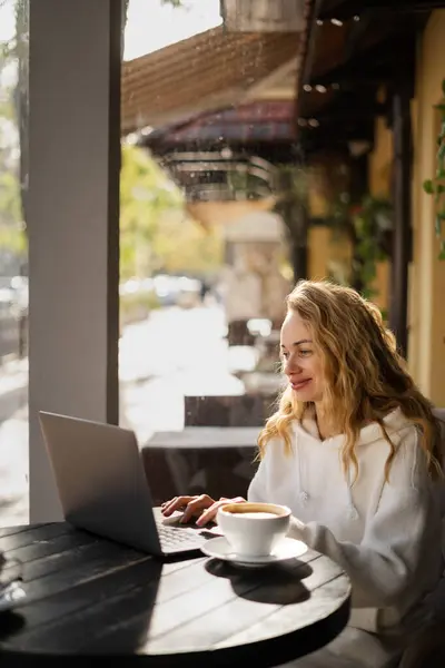 Blonde woman typing and watching on laptop computer in cafe sitting at table near window. Freelance and remote work concept
