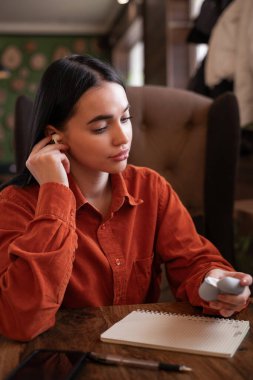 Bayan öğrenci kahve evinde otururken kulaklığına kablosuz kulaklık takıyor. Kadın eli, kablosuz bluetooth kulaklık kutusu tutuyor. Boşluğu kopyala