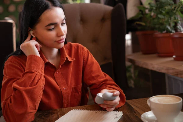 Time to listen to music and relax concept. Cute young woman takes out modern mini wireless bluetooth earphones from a charging case and put into ear. Cup of coffee on the table. Copy space
