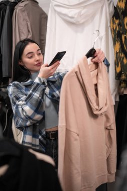 Kadın müşteri elbise mağazasından alışveriş yapıyor, akıllı telefonuyla fotoğraf çekiyor, stil dükkanından şık kıyafetler seçiyor. Boşluğu kopyala