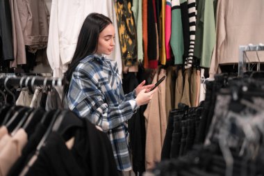 Giyim mağazasından alışveriş yapan güzel bir bayan alıcı akıllı telefonunu kullanarak internetten şık kıyafetler seçiyor. Moda Dükkanı konsepti