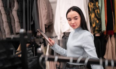 Genç esmer satıcı galeride yeni kıyafet koleksiyonlarıyla dikilirken dijital tablet kullanıyor. Boşluğu kopyala