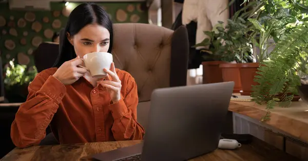 Beautiful woman drinking coffee and working on her laptop in a coffee shop at wooden table. Copy space