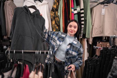 Giyim mağazasında kazak tutan genç müşteri kadın üzgün bir ifadeyle seçim için endişeleniyor. Boşluğu kopyala.