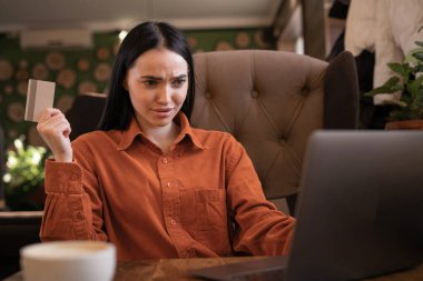 Kredi kartıyla kafası karışmış genç bir kadın çevrimiçi sipariş ödemeleriyle ilgili düşünceli bir şekilde kafa yoruyor, banka hesabına internetten alışveriş yapmasını engelliyor kafede dizüstü bilgisayar kullanıyor. Boşluğu kopyala