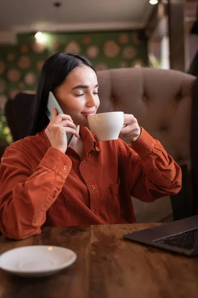 Young woman talking with client about new project on Mobile phone call. Brunette girl chatting with friend by smartphone while sitting in a coffee shop. Drinking coffee in cafe