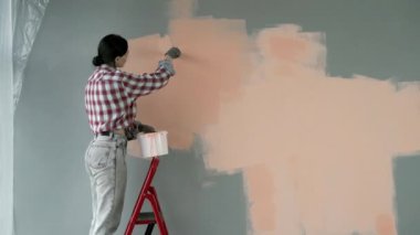 Kadın ressam, merdiven üzerinde dururken fırçasıyla bej bir duvar boyası boyuyor. Ev yenileme konsepti
