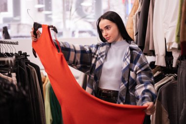Mutlu esmer kadın alışveriş merkezinde ya da giyim mağazasında elbise seçiyor ve elinde turuncu elbiseler tutuyor. Alışveriş, moda, tarz ve insan konsepti. Boşluğu kopyala
