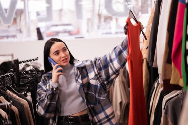 Genç bayan müşteriler giyim mağazasından alışveriş yapıyor, şık elbiseler seçiyor, akıllı telefon kullanıyor ve onu arıyor. Moda Dükkanı. Satış, moda, tüketim ve insan konsepti. Boşluğu kopyala