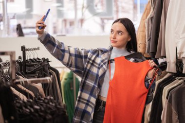 Alışveriş, moda, tarz, teknoloji ve insan konsepti. Akıllı telefonlu genç esmer kadın yeni turuncu elbisesiyle elbise mağazasında selfie çekiyor. Boşluğu kopyala