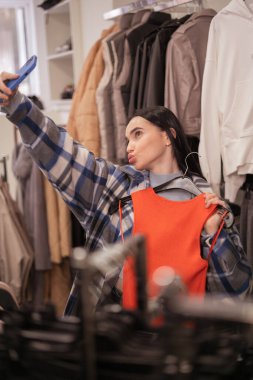 Alışveriş, moda, tarz, teknoloji ve insan konsepti. Akıllı telefonlu genç esmer kadın yeni turuncu elbisesiyle elbise mağazasında selfie çekiyor. Boşluğu kopyala