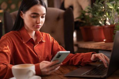 Kadın serbest çalışan akıllı telefonlara göz atıyor ve açık bir kafede oturup internette dizüstü bilgisayarda çalışıyor. Kafeteryada blog yazarı. Boşluğu kopyala