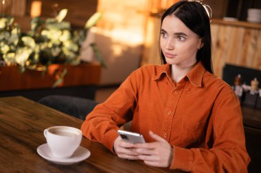 Kahve içen, kafede cep telefonu kullanan ve dinlenirken cep telefonundan iyi haberler okuyan tatlı, genç esmer kadın. Boşluğu kopyala