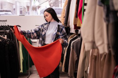 Alışveriş merkezinde ya da giyim mağazasında kıyafet seçen ve turuncu elbiseye bakan genç bir kadın. Alışveriş, moda, tarz ve insan konsepti. Boşluğu kopyala