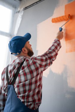 Mavi tulumlu ve şapkalı yakışıklı ressamın boya silindiriyle duvarı boyadığını gördüm. Yenileme, dekorasyon ve onarım konsepti.