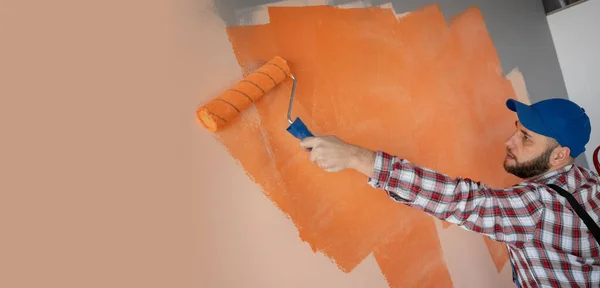 Painter in blue cap and overalls painting a wall with paint roller. Builder worker painting surface with orange color. Banner. Copy space