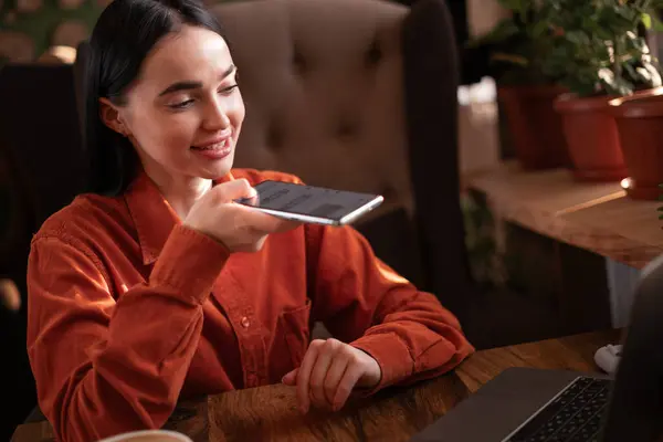 Young woman inside cafe working with laptop, recording audio message using app on smartphone, girl using artificial intelligence assistant. Copy space