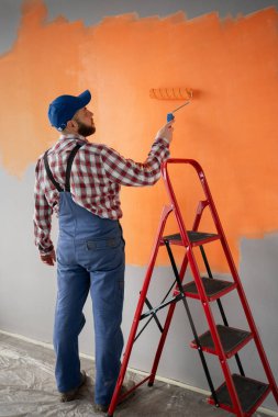 Ressamın turuncu renkli, boya silindirli ve kırmızı merdiveni olan boş bir duvarı boyamasının arka görüntüsü. Boşluğu kopyala