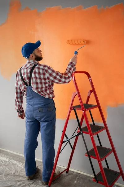 Rear view of painter man painting blank wall in orange color, with paint roller and red ladder. Copy space