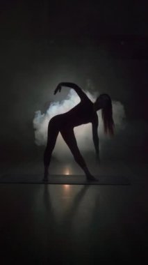 Karanlık bir salonda dumanla yoga yapan genç bir kadının silueti. Yoga alıştırması konsepti