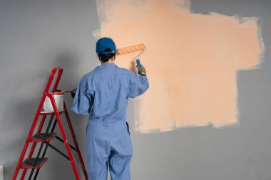 Mavi tulumlu, kepli ve eldivenli boyalı duvarı boyayan genç kadın ressamın arka görüntüsü. Boşluğu kopyala