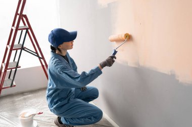 Önlüklü ve eldivenli kadın ressamın arka planda boya silindiri, merdiven ve kovalarla duvarı boyadığını gördüm. Boşluğu kopyala