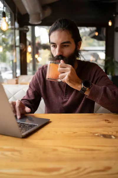 Portrait of bearded man working remotely on laptop computer in cafe interior, hipster guy drinking tea while updating software. Copy space