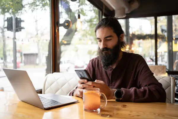Young bearded hipster man answering the client by phone while having break in a coffee shop. Long haired guy searching the information typed in a mobile phone and laptop. Copy space