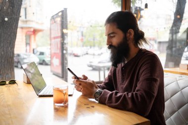 Kafede çalışan genç adam akıllı telefon ve dizüstü bilgisayar kullanıyor, hippi adam dahili kahve dükkanında akıllı telefon kullanıyor, işyerinde teknoloji kullanıyor. Boşluğu kopyala