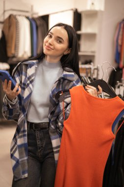 Giyim mağazasında akıllı telefonu olan mutlu esmer bir kadın. Alışveriş, moda, tarz, teknoloji ve insan kavramı