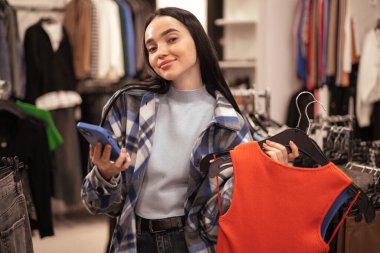 Giyim mağazasında, moda mağazasında ya da alışveriş merkezinde beklerken elinde parlak elbiseler ve akıllı telefonlar tutan mutlu genç bayan alışverişçi. Satış, alışveriş ve insanlar konsepti