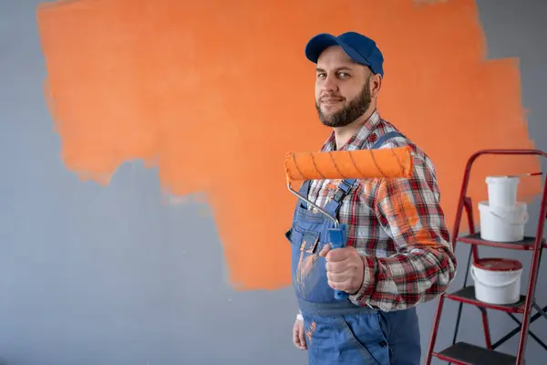 Portrait of one male house painter worker in blue overalls and cap with painting roller before orange wall. Copy space