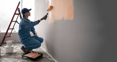 Önlüklü ve eldivenli kadın ressam arka planda boya rulosu, merdiven ve kovalarla bir duvar boyuyor. Yan görüş. Boşluğu kopyala