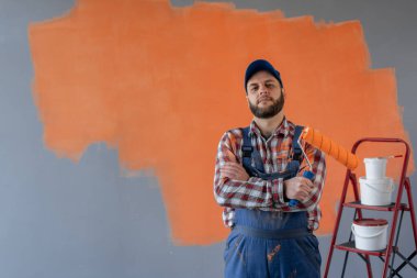 Çapraz kollu boyalı ressam elinde boya silindiriyle kameraya bakıyor, doğal bir yüzü ve kendine güvenen bir ifadesi var. Boşluğu kopyala
