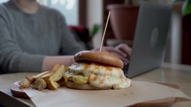おいしいハンバーガーやチーズバーガーは リモートでラップトップ 選択的なフォーカスで作業するクライアントフリーランサーにカフェでフライドポテトを提供しました クローズアップ — ストック動画